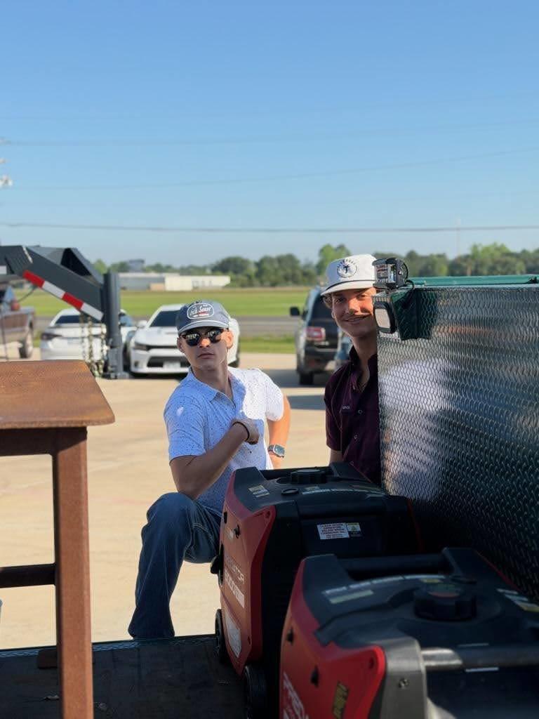 Two people sit by a table, one wearing a hat and sunglasses, the other with a camera. Behind them, a vehicle with a flatbed trailer is parked.
