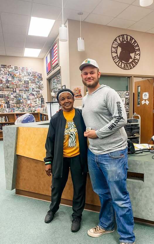 Two people stand in a room. One wears a yellow shirt, and the other wears a gray sweatshirt. They stand near a wooden desk.