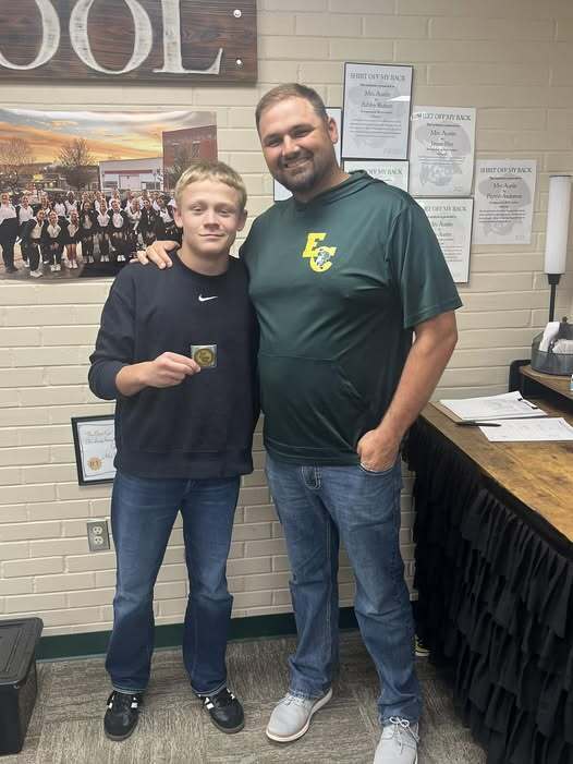 Two men stand together; one holds a medal and they both smile. Behind them, a wall displays school posters and a desk with papers.