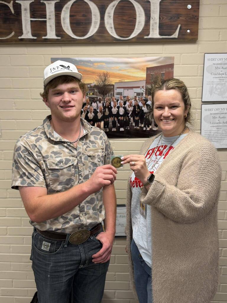 A man and a woman pose with a medal in front of a brick wall displaying a "School" sign and other framed certificates.