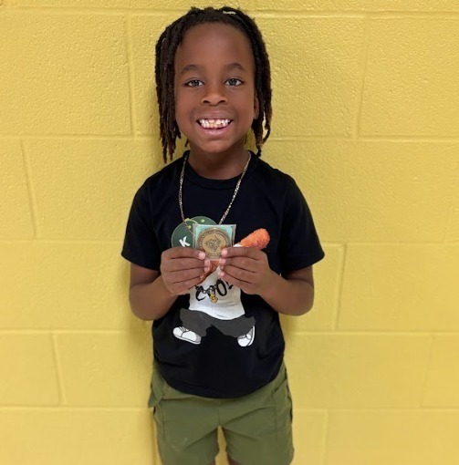 A young child with braided hair stands before a yellow wall, holding a carrot and a card.
