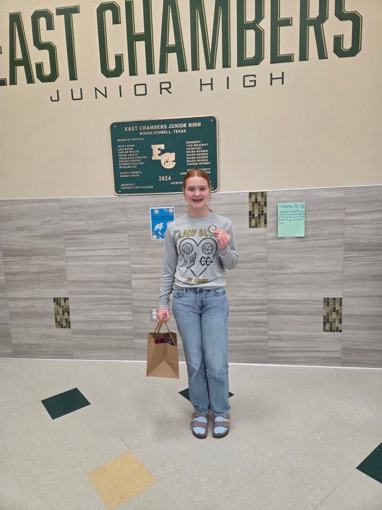 A girl stands in a hallway with a sign behind her reading "EAST CHAMBERS JUNIOR HIGH". She holds a brown bag.