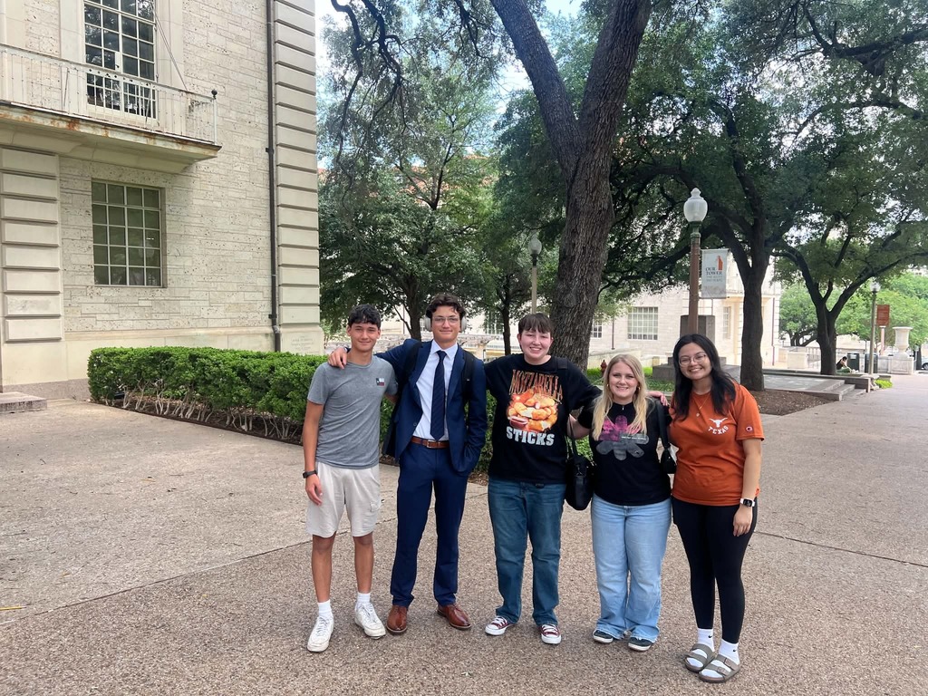 Five people pose in front of a building, wearing casual attire. Behind them are shrubs and trees.