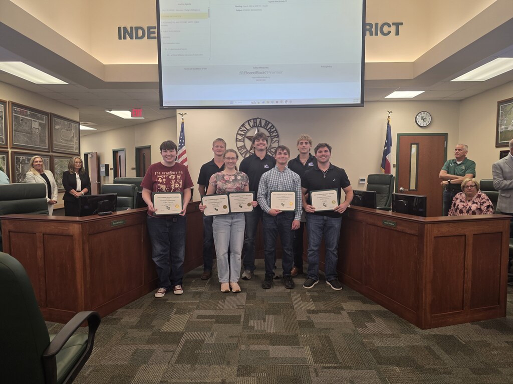 A group of people stand holding certificates in a council chamber, with two flags, a projector screen, and a clock.