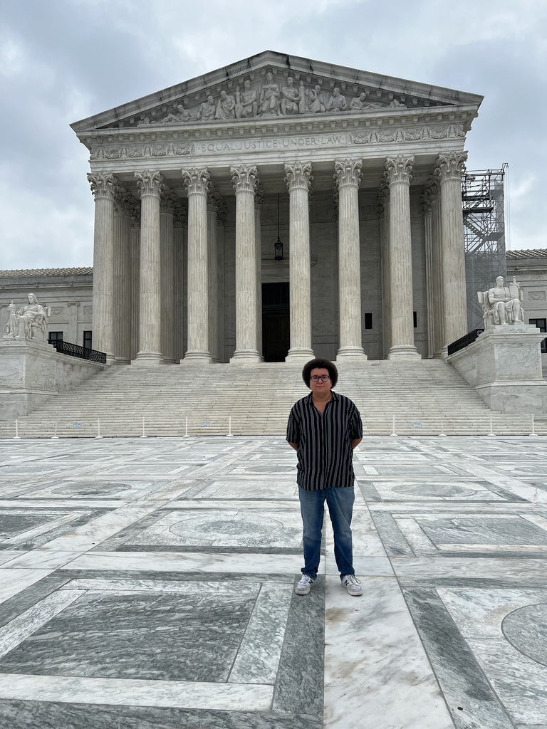 Person stands in front of the Supreme Court building. The building has tall columns, and steps lead up to the entrance.