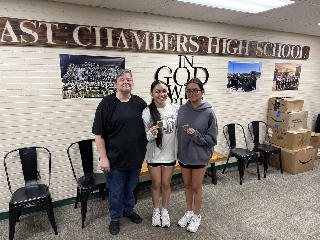 Three people stand next to each other in a room with a "Last Chambers High School" sign. A man and two girls.
