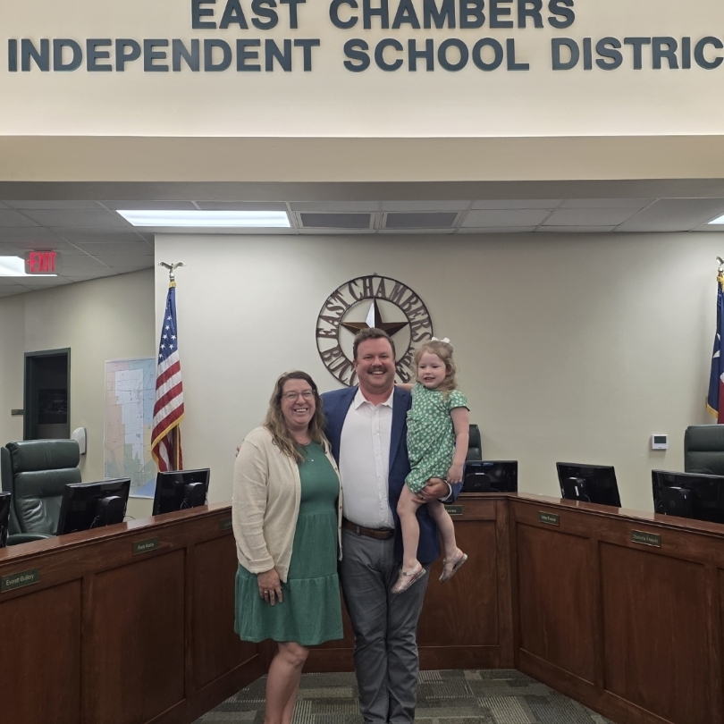 Two adults and a child stand in front of a podium in a conference room. Flags hang behind them.