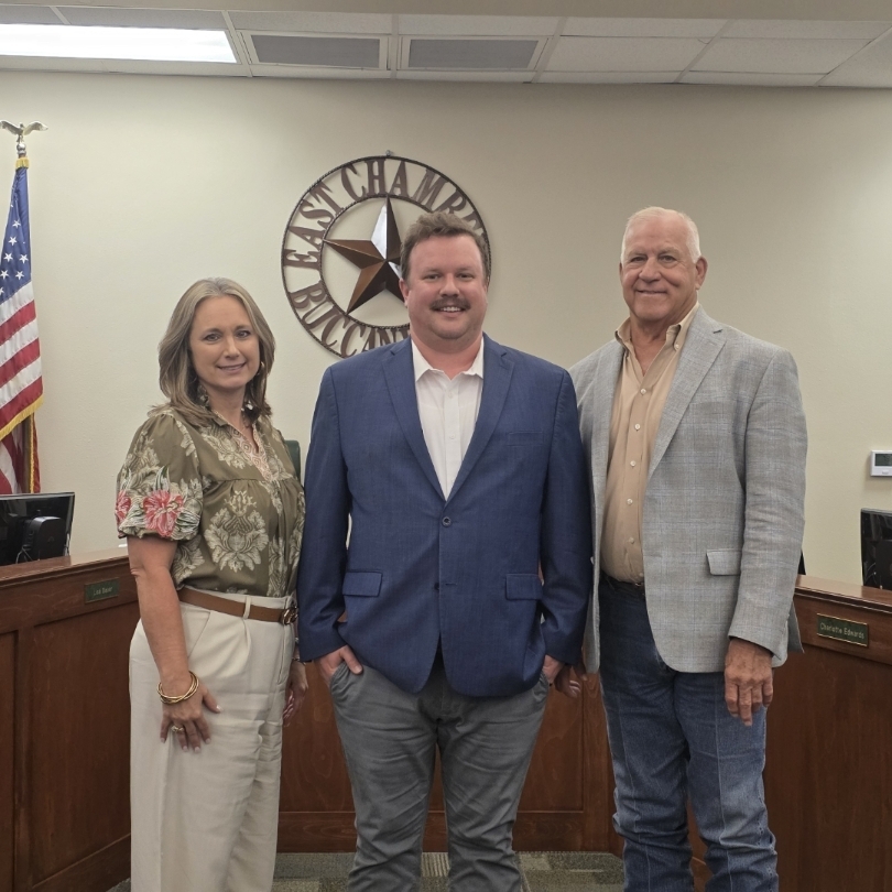 Three people in formal wear standing in front of a city council meeting table, with flags and a sign.