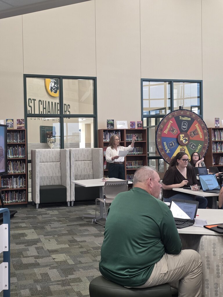 Library with patrons seated at tables. A woman stands behind a partition and speaks to a group.