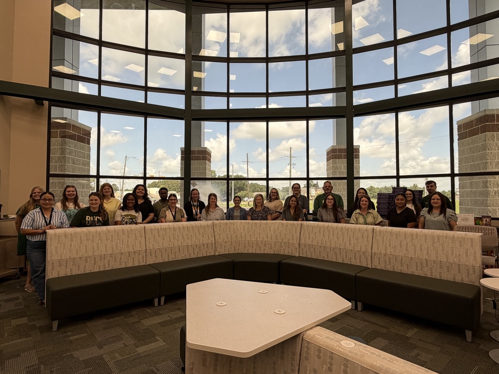 Group of people seated in a semicircle around a table in a room with large windows.
