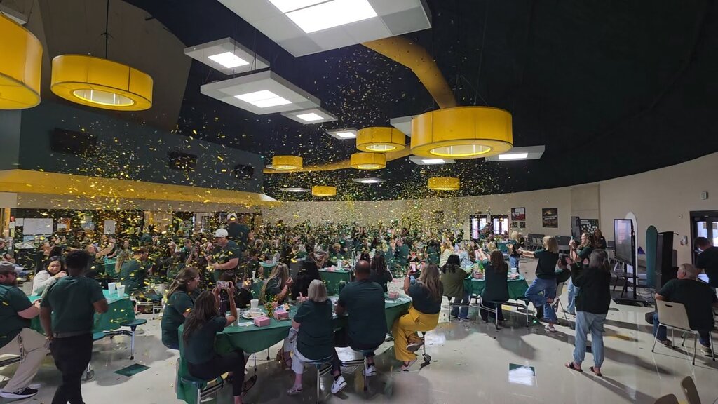 Large indoor event space with seated tables. Participants wearing green shirts. Confetti falling from the ceiling.