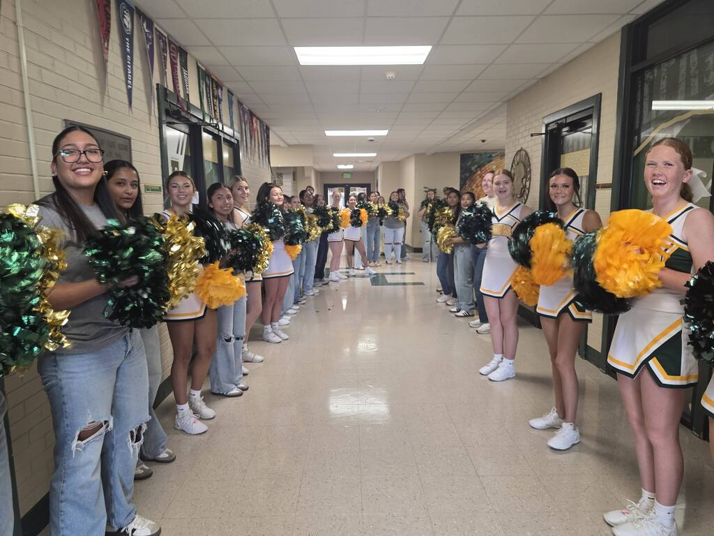 Cheerleaders in uniform with yellow and green pom-poms line the hallway, smiling and posing.