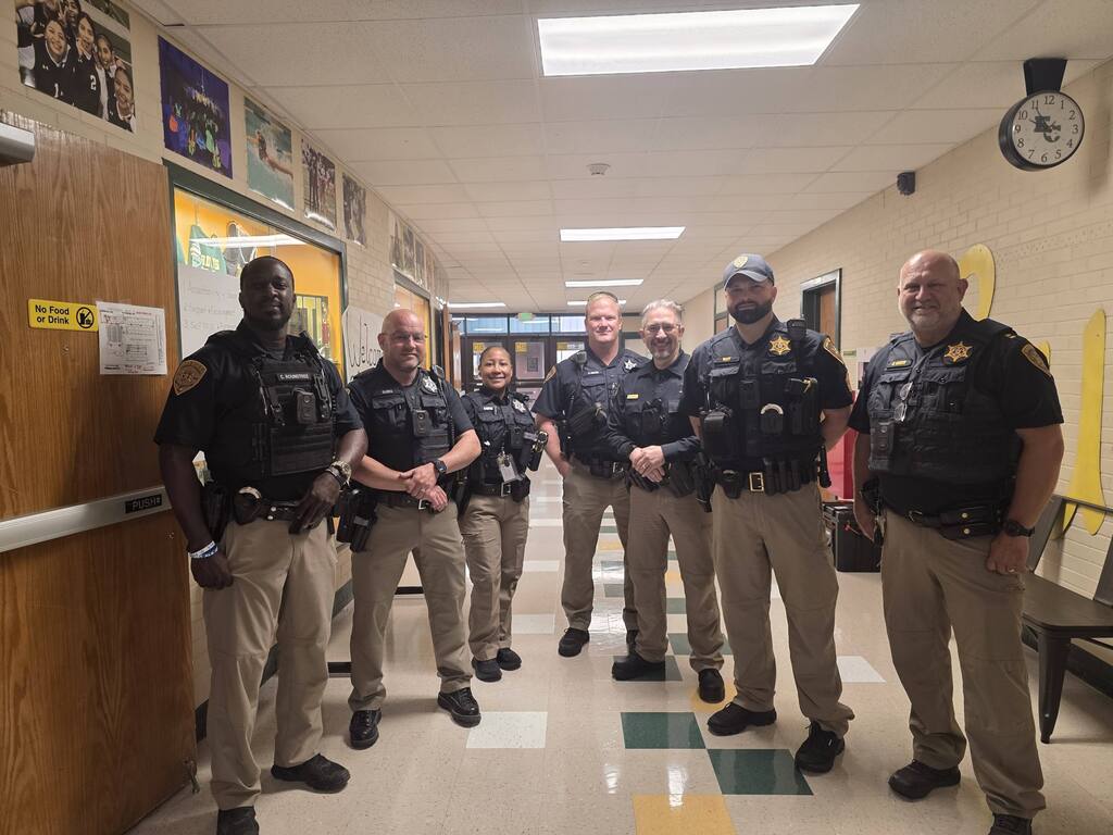 Six police officers stand in a hallway, two with hands on hips. One officer wears a hat.