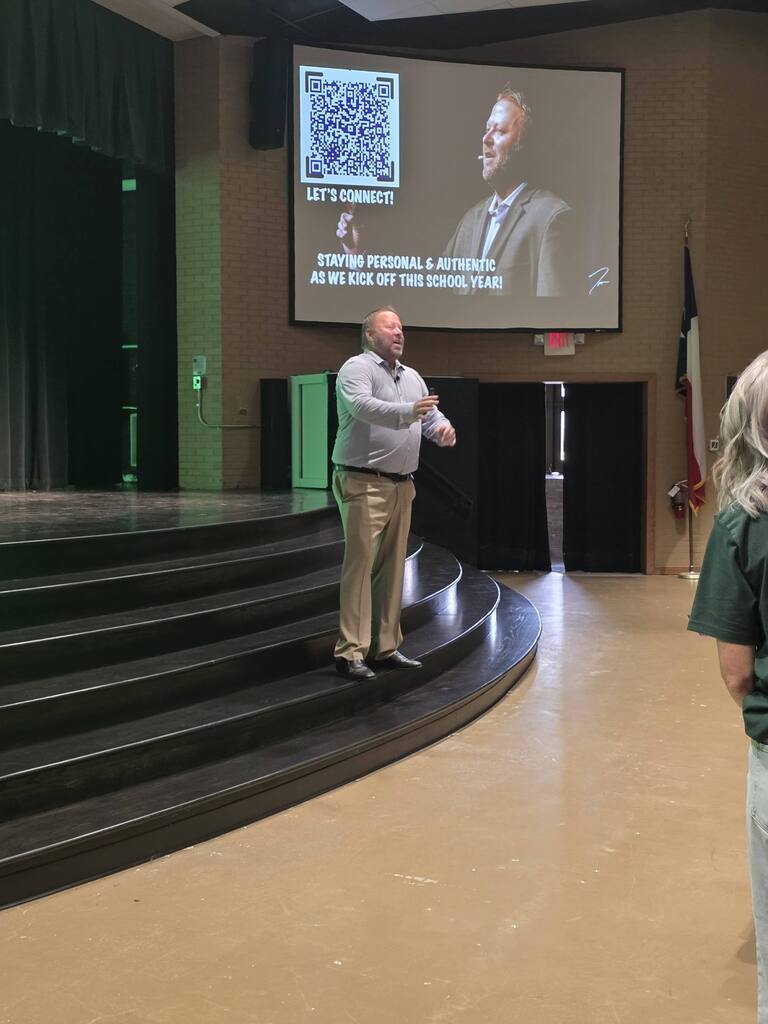 A man stands on a stage, facing an audience, with a large screen behind him displaying an image of another man.