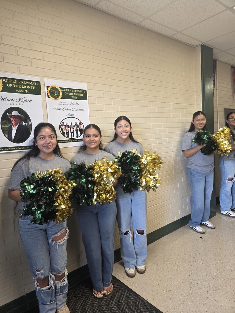 Five cheerleaders in blue uniforms and jeans hold gold and green pom-poms, standing against a wall with posters.
