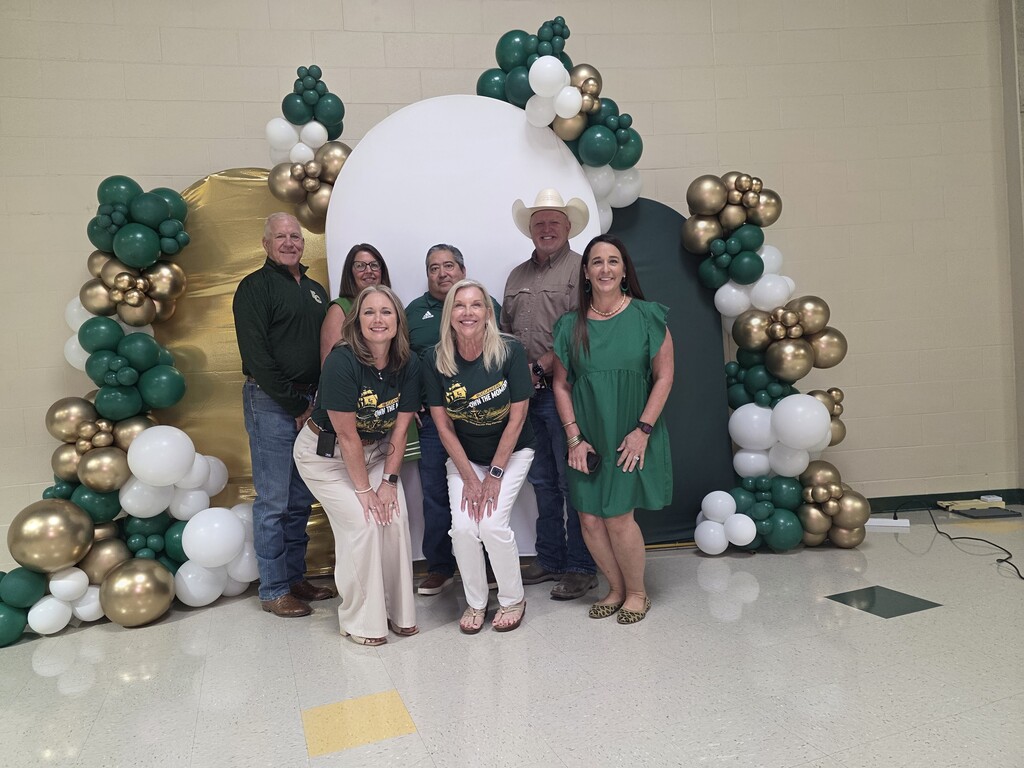 A group of people in green shirts standing in front of a decorated archway with gold and green balloons.