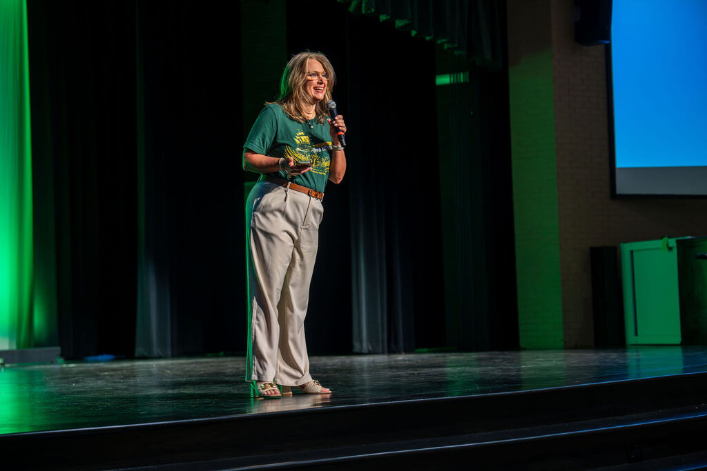 A woman on stage with a microphone, green light illuminating the background. She wears a green shirt and white pants.