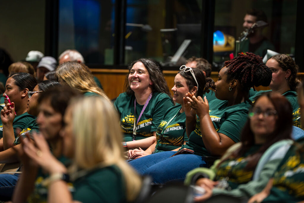 A room with people seated, mostly women, wearing green shirts with yellow accents, clapping hands and smiling.