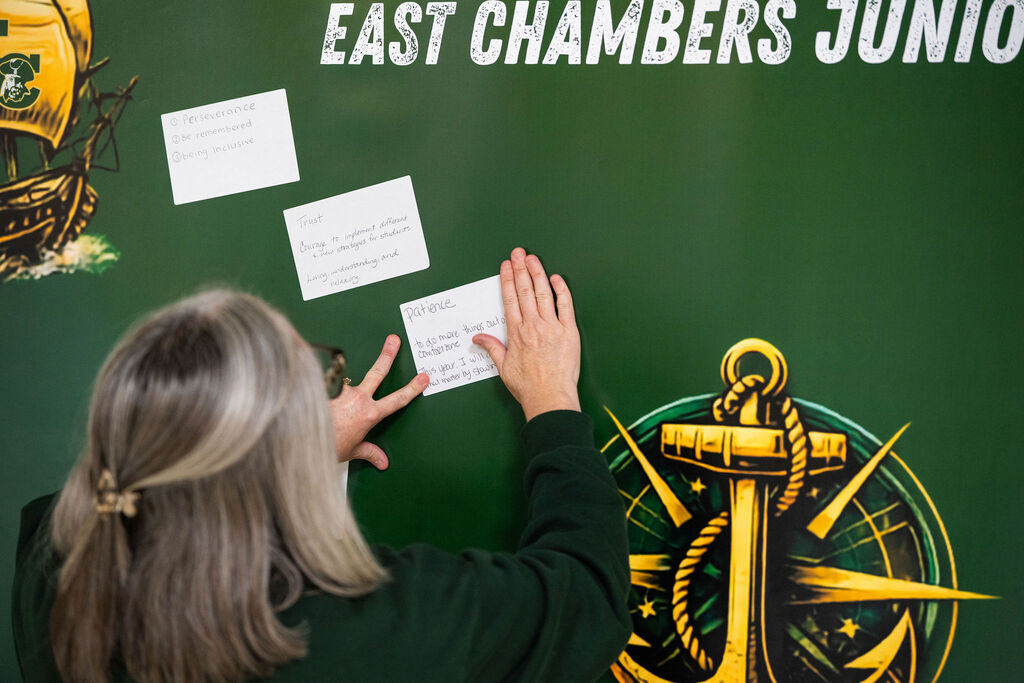 An adult places a paper on a bulletin board that reads "EAST CHAMBERS JUNIO" with three other papers.