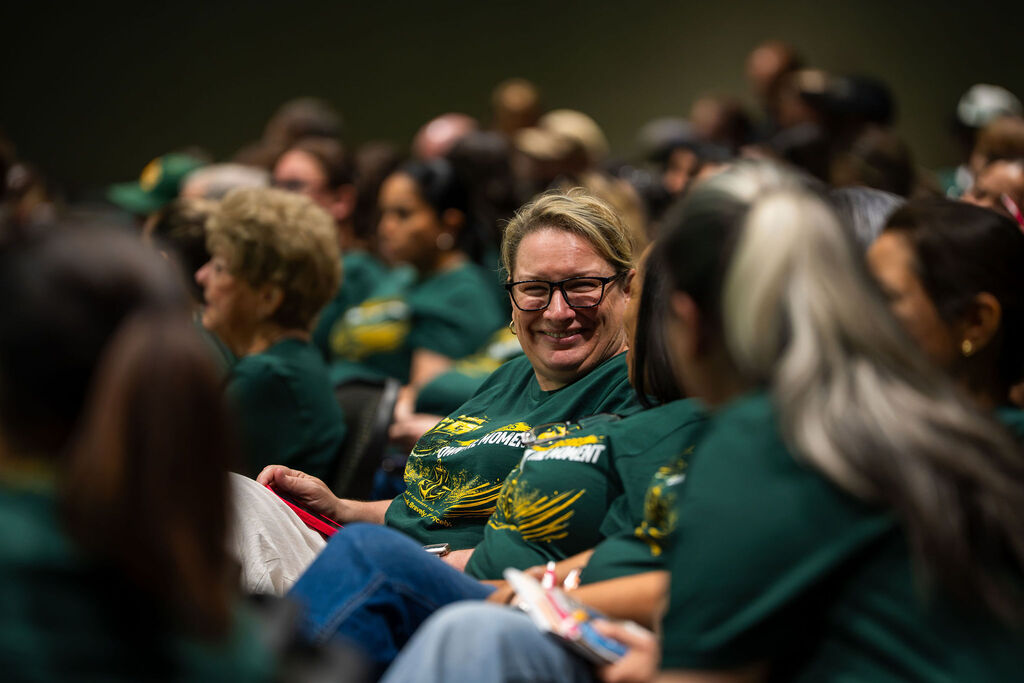 Several people wearing green shirts and glasses are seated in an auditorium. One person smiles at the camera.