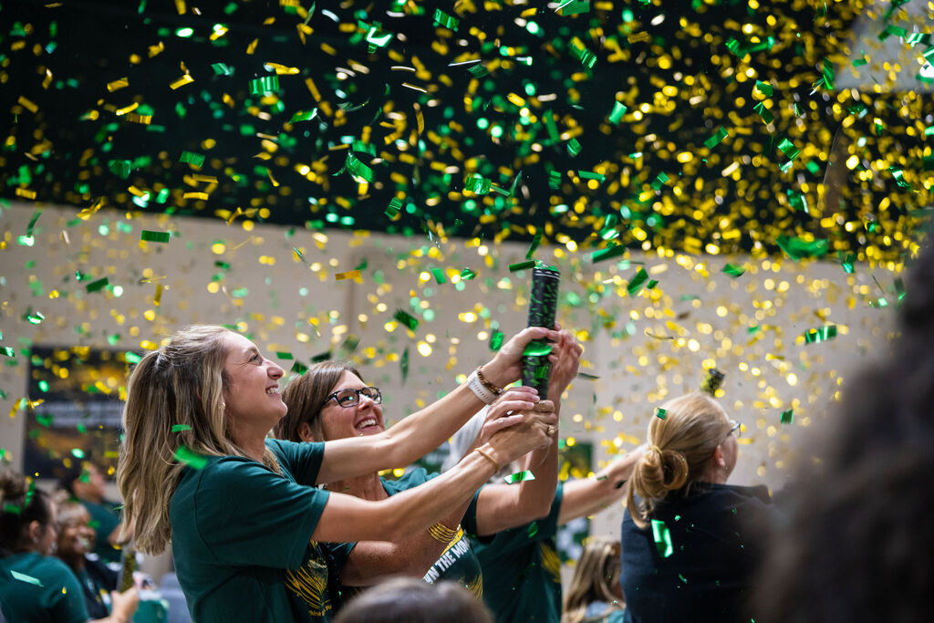 A group of people in green shirts inside a hall, holding up a green object and celebrating.