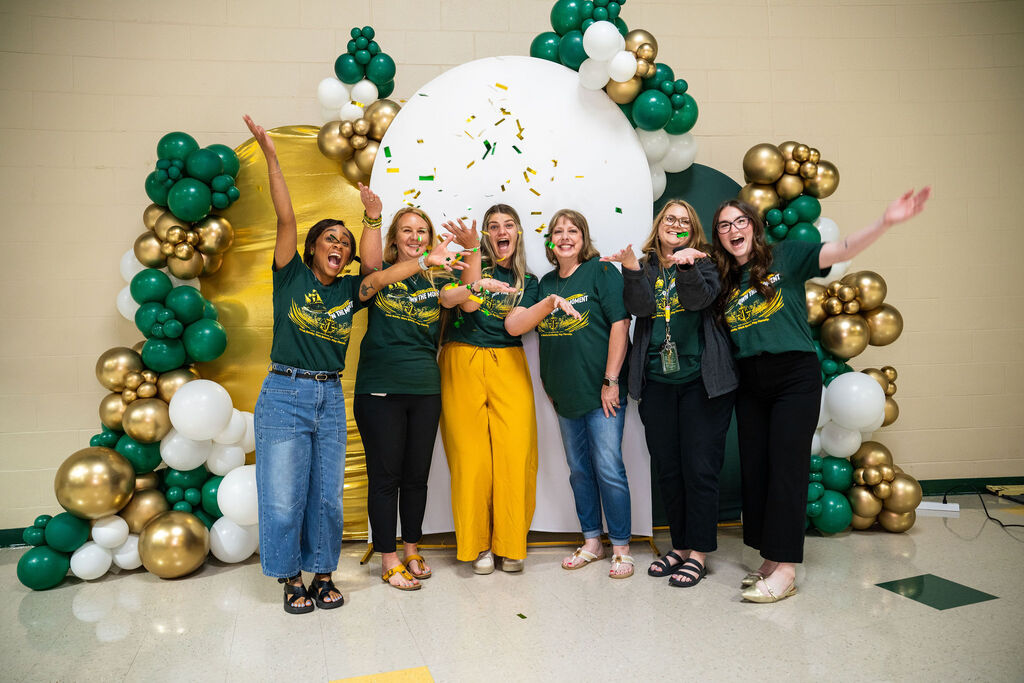 A group of women, all in matching green shirts, pose with raised hands in front of a colorful arch.