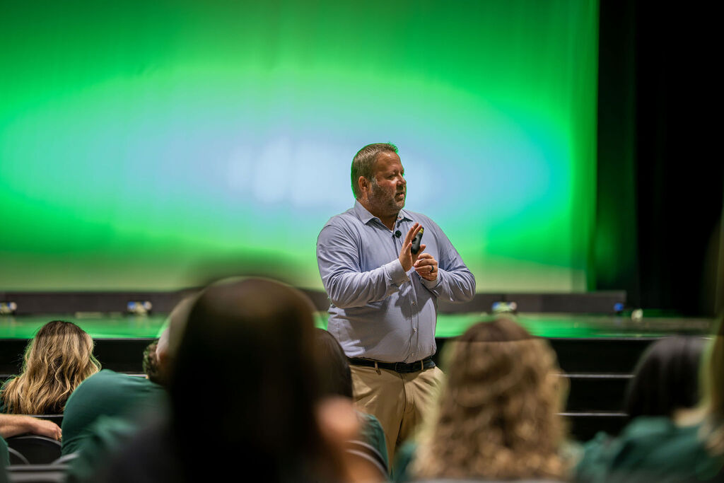 A man speaks in front of a green screen while addressing a seated audience in a room.