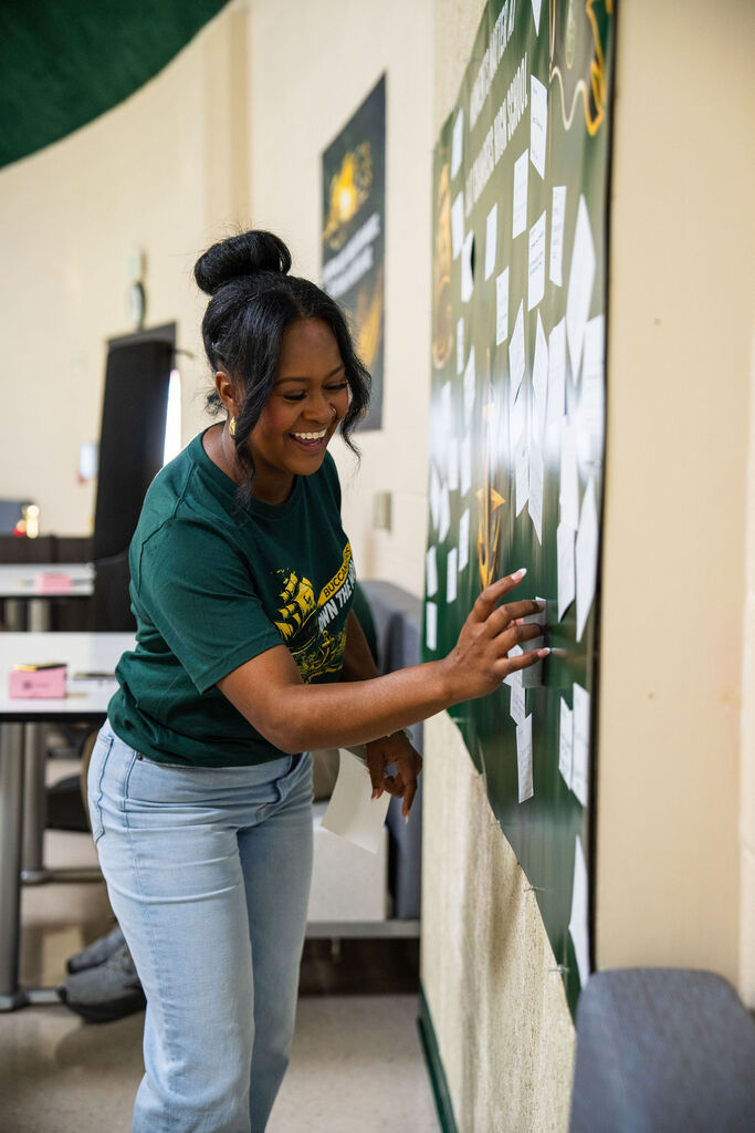 Woman in green T-shirt standing near bulletin board, placing paper with hands, smiling.