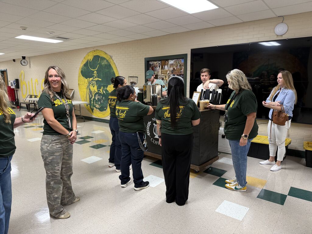 A group of people in matching green shirts stand around a counter in a room with a yellow wall and checkered floor.