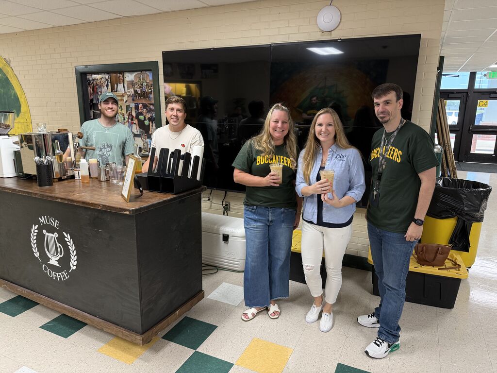 Five people stand behind a small counter with bottles, glasses, and cups. A large monitor is behind them.