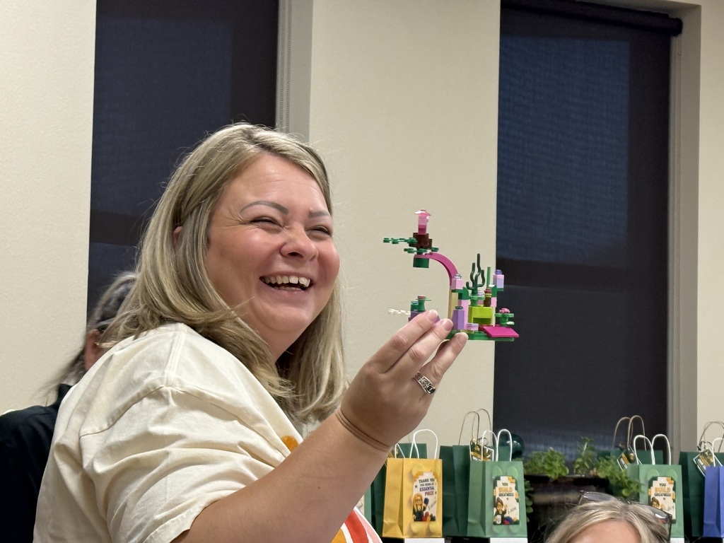 Woman with blond hair smiling while holding a small multi-colored Lego creation, with several gift bags in the background.