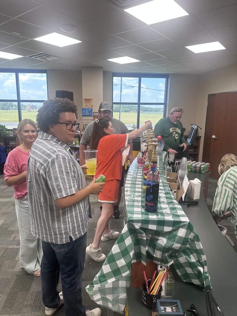 A group of people are standing around a long table with various items on it, including bottles and cups.