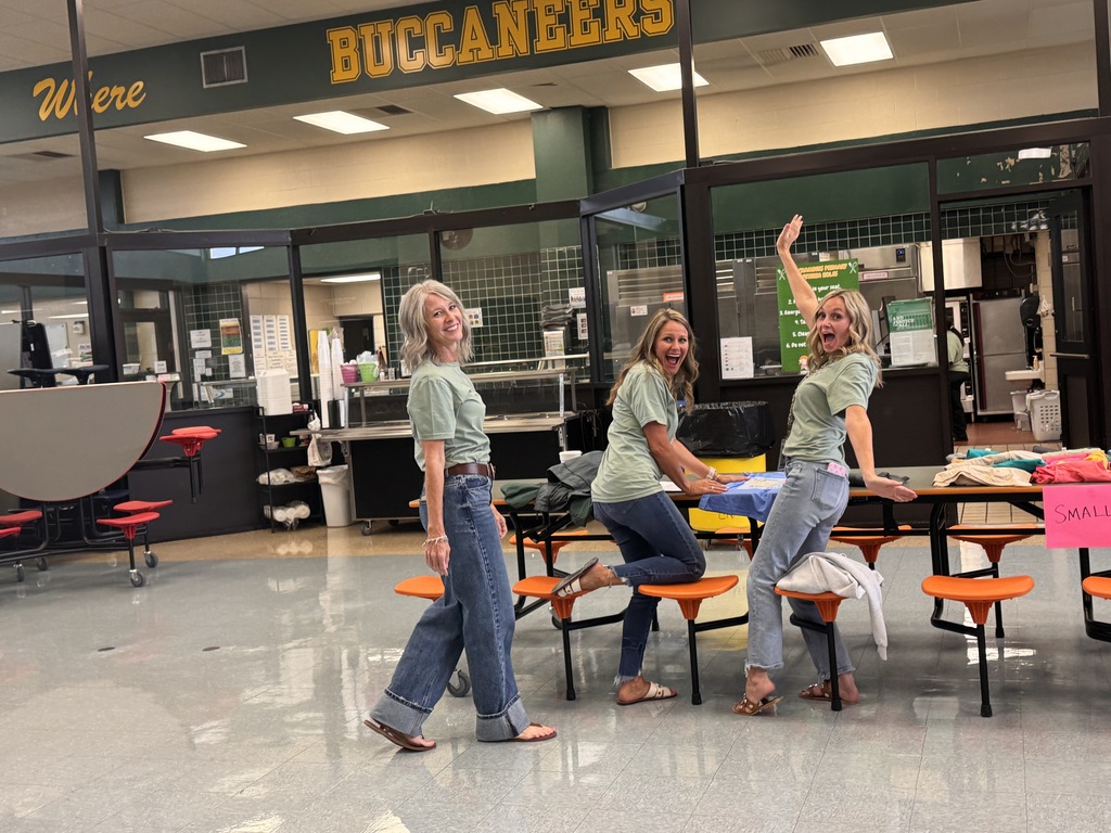 Three women in green shirts, posing with arms raised, at a table in a cafeteria. Behind them, a sign reads "BUCCANEERS" above a glass entrance.