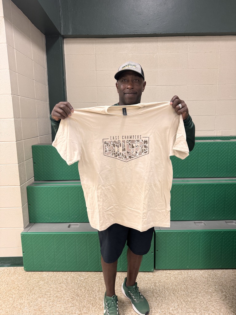Man in cap and sneakers holds up a white T-shirt with black text in a room with green bleachers.