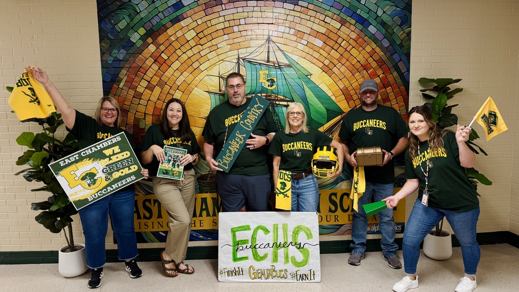Group of people in green shirts pose with signs and flags in a room with a large mural.