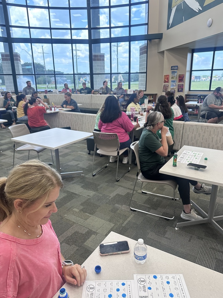 Room with many people sitting at tables playing a game. One woman in pink sits at a table.