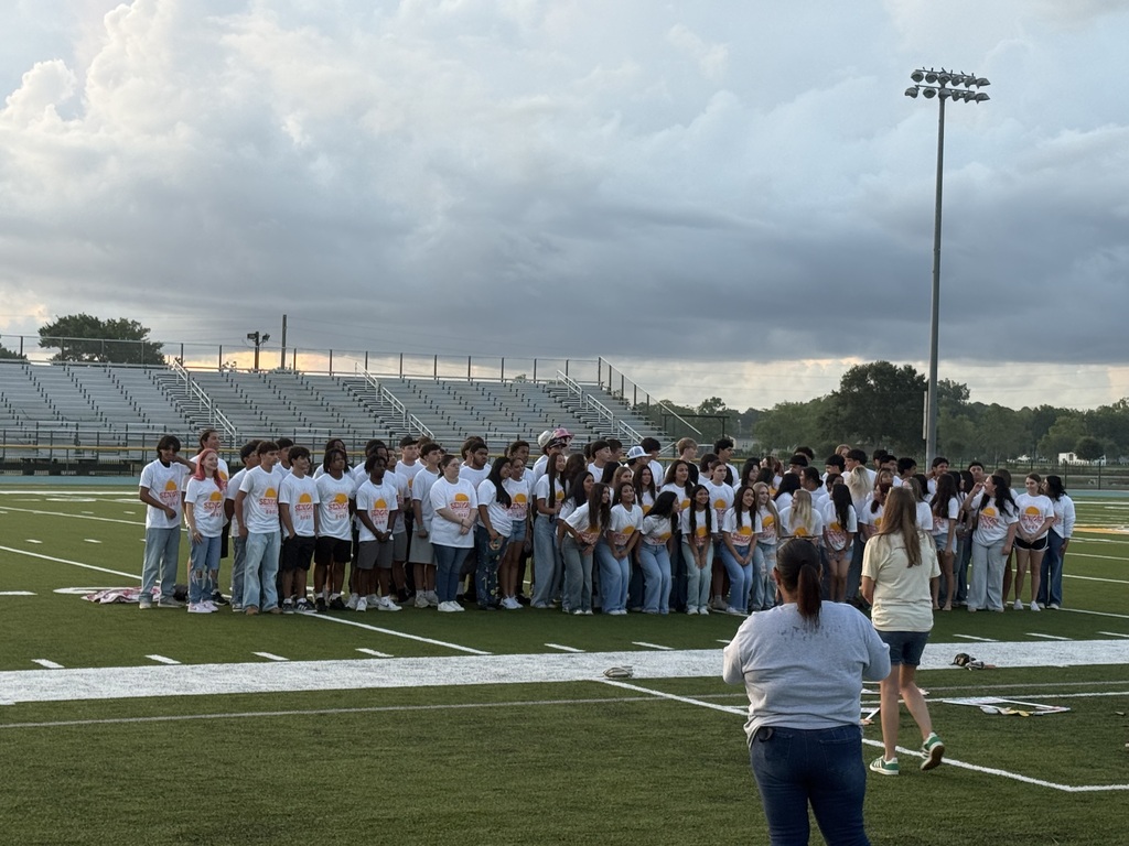 A group of people in white shirts and jeans stands on a football field. A person in blue jeans and a gray shirt stands at the front.