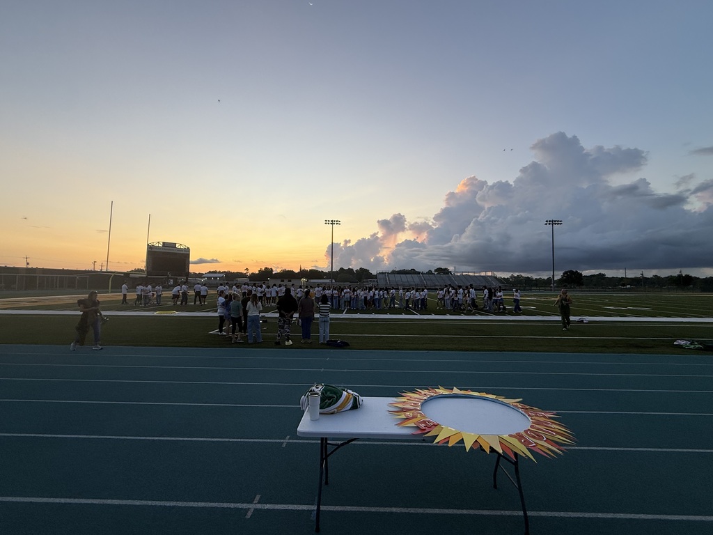 Track field with a sunset sky. A table has a circular decoration. Many people are standing in groups.