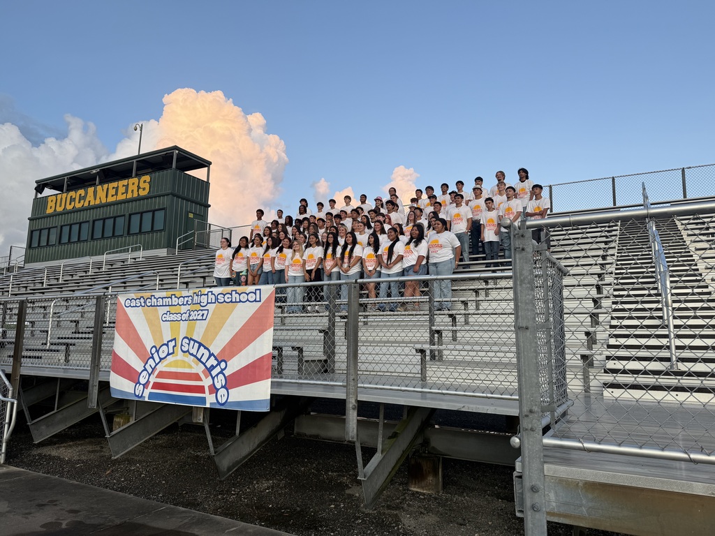 A group of people in white shirts stand on a stadium's upper tier, facing the camera. A banner is behind them.