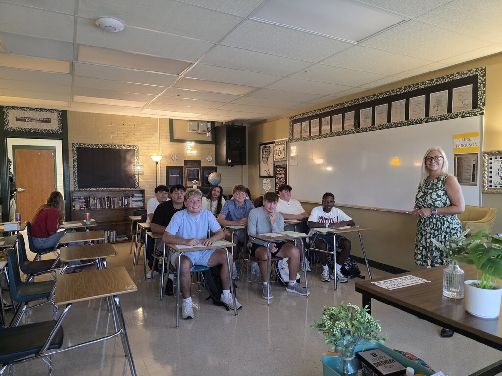 A classroom with students seated in chairs around desks, a woman stands in front of them, next to a white board.