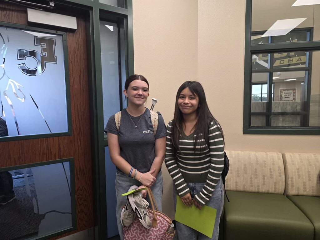 Two women standing in a hall, one holding a folder, other with a bag, next to a bench.