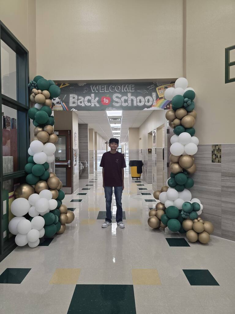A person stands in a hallway under a balloon arch that reads "Welcome Back School". The arch is green and white.