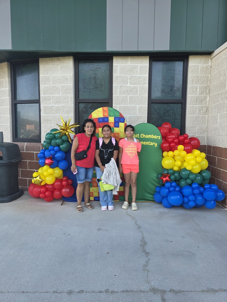 Three children stand in front of colorful balloon decorations and a building with windows.