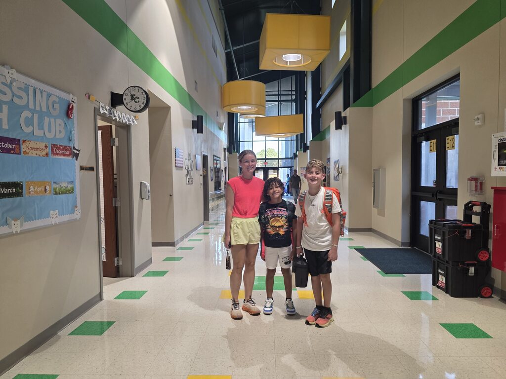 Three children stand in a school hallway. A bulletin board is on the left, and a door is in the background.
