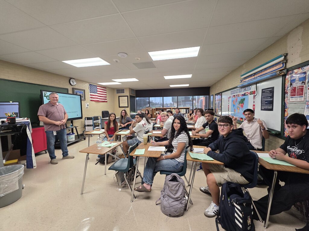 A classroom with desks and chairs; a teacher stands at the front with a screen. Students sit in rows, engaged in class.