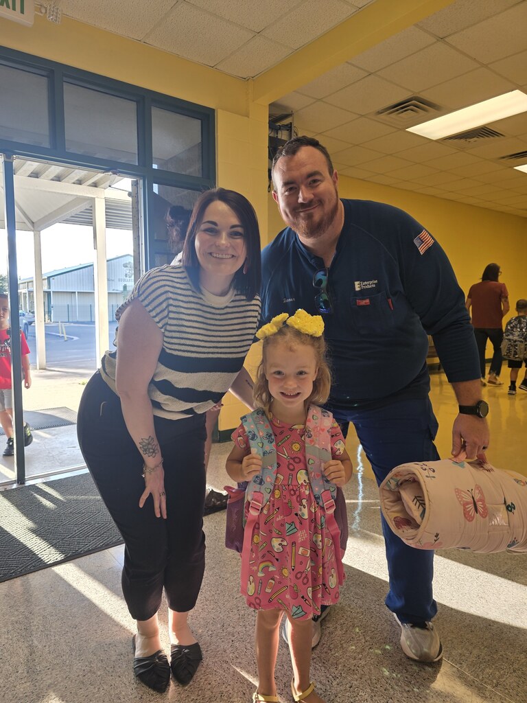 A woman, man, and young girl pose in a yellow hallway. The man holds a rolled item, and the girl wears a floral dress.