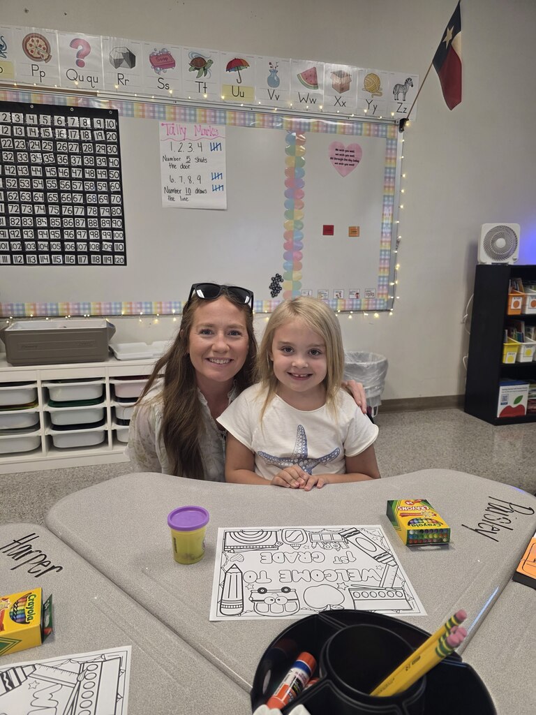 A woman and a young girl sit at a table in a classroom, surrounded by art supplies.
