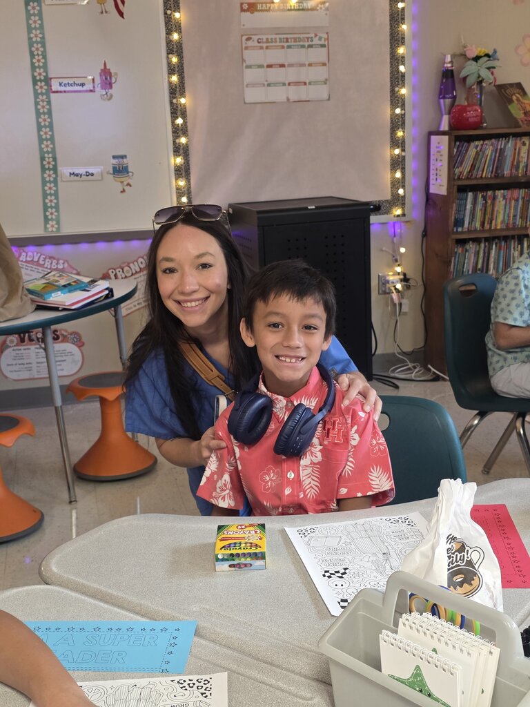 Woman with sunglasses, blue shirt, hugging a smiling boy in a pink shirt, at a desk.