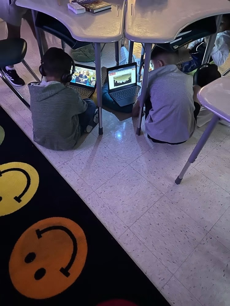 Two children sit under desks, using laptops. One wears headphones. A black rug with smiley faces is on the floor.