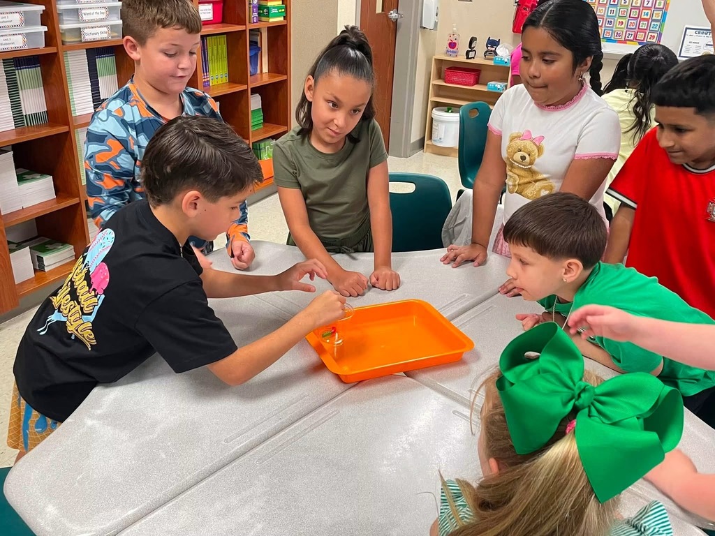 Group of children seated around a table in a classroom. One child is pouring sand into an orange tray.